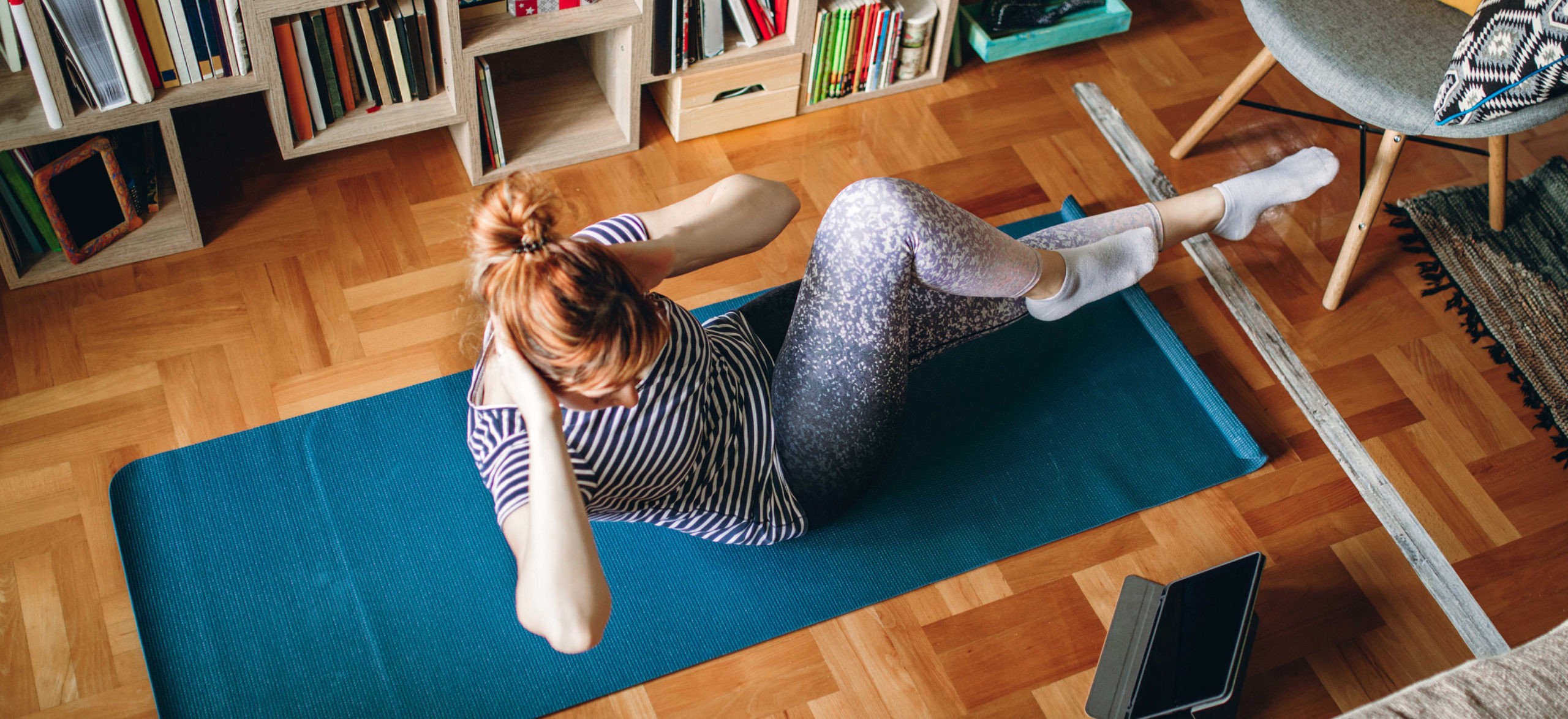 Woman in striped shirt and tights exercising on yoga mat while looking at tablet.
