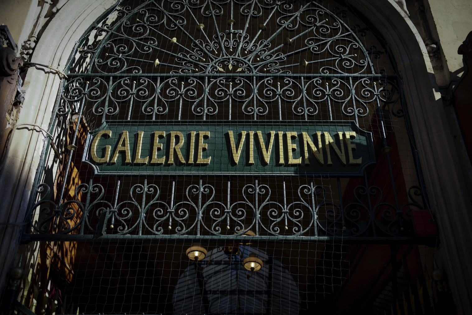 A beautiful metal shop sign that reads 'Galerie Vivienne' is splattered with shadows in the afternoon sun.