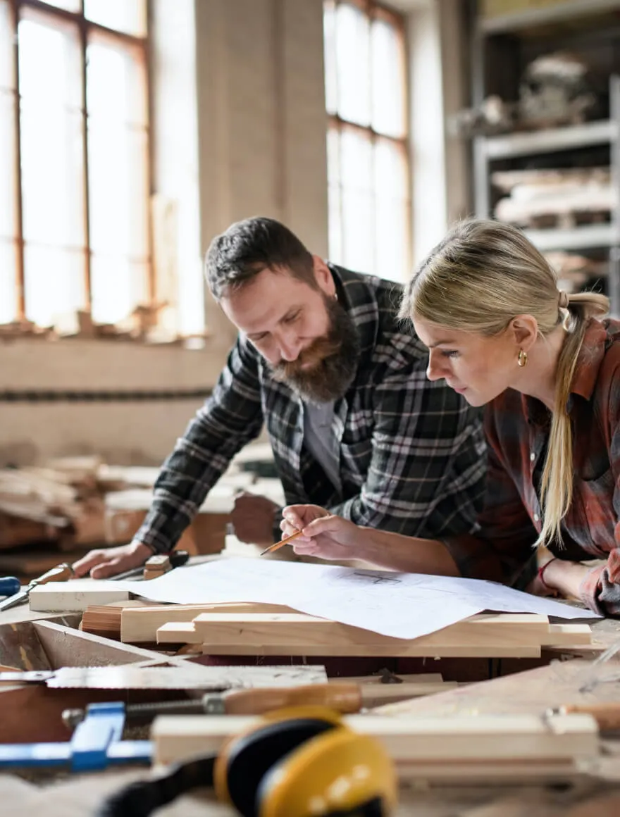 woman and bearded man wearing flannels leaning over work table looking at blueprint on top of wood pieces and tools