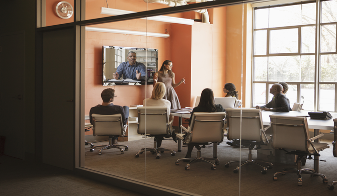 conference room with several participants watching a woman present and one remote participant on video conference