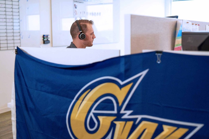Man sitting in cubicle wearing wireless headphones next to a George Washington University banner