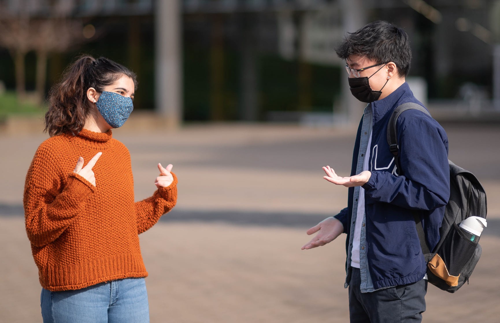 Two people wearing COVID-19 face masks use hand gestures to help communicate to each other