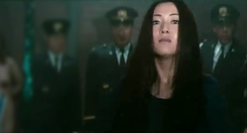 A screenshot of a woman standing off-center from the camera with her chin up as guards loom menacingly behind her. From the movie 'Female Prisoner 701: Scorpion'.