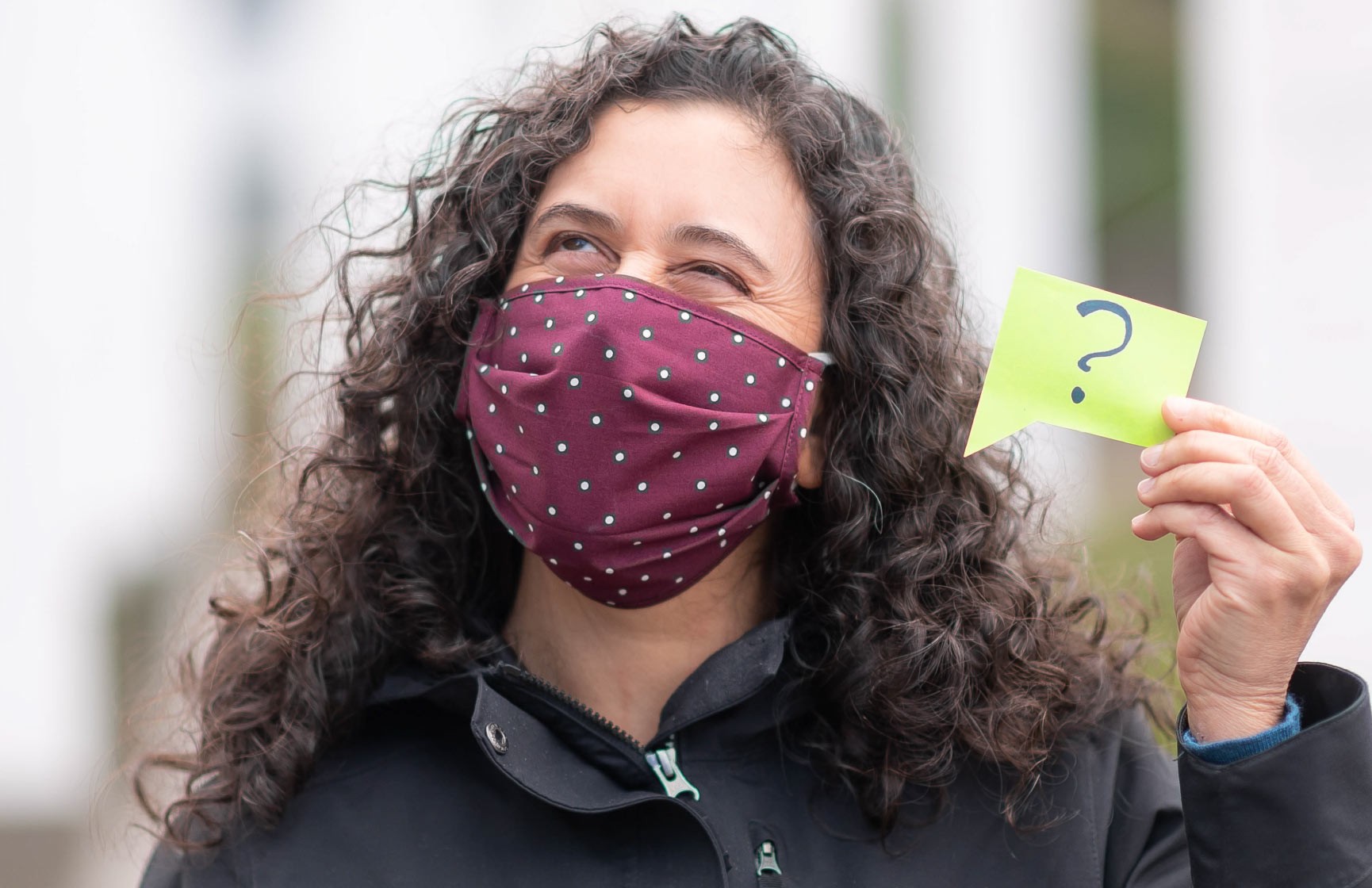 A person wearing a COVID-19 face mask holds up a sign with a question mark on it to indicate that they can't understand what people are saying