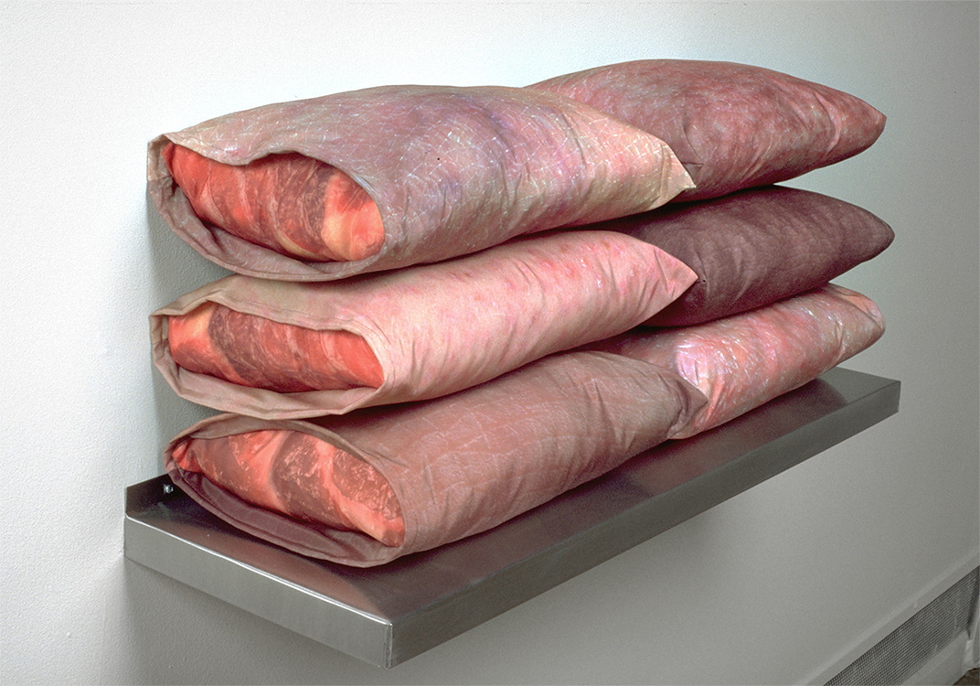 Six pillows, stacked in sets of three, side by side, sit on a metal shelf on a wall. The pillow cases and inner pillow material have the color and marbling of raw meat.