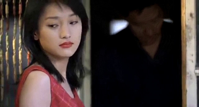 A close-up screenshot from the movie 'Beijing Bicycle' of a young woman with red lipstick in a red qipao dress shifting her eyes off camera to the left.