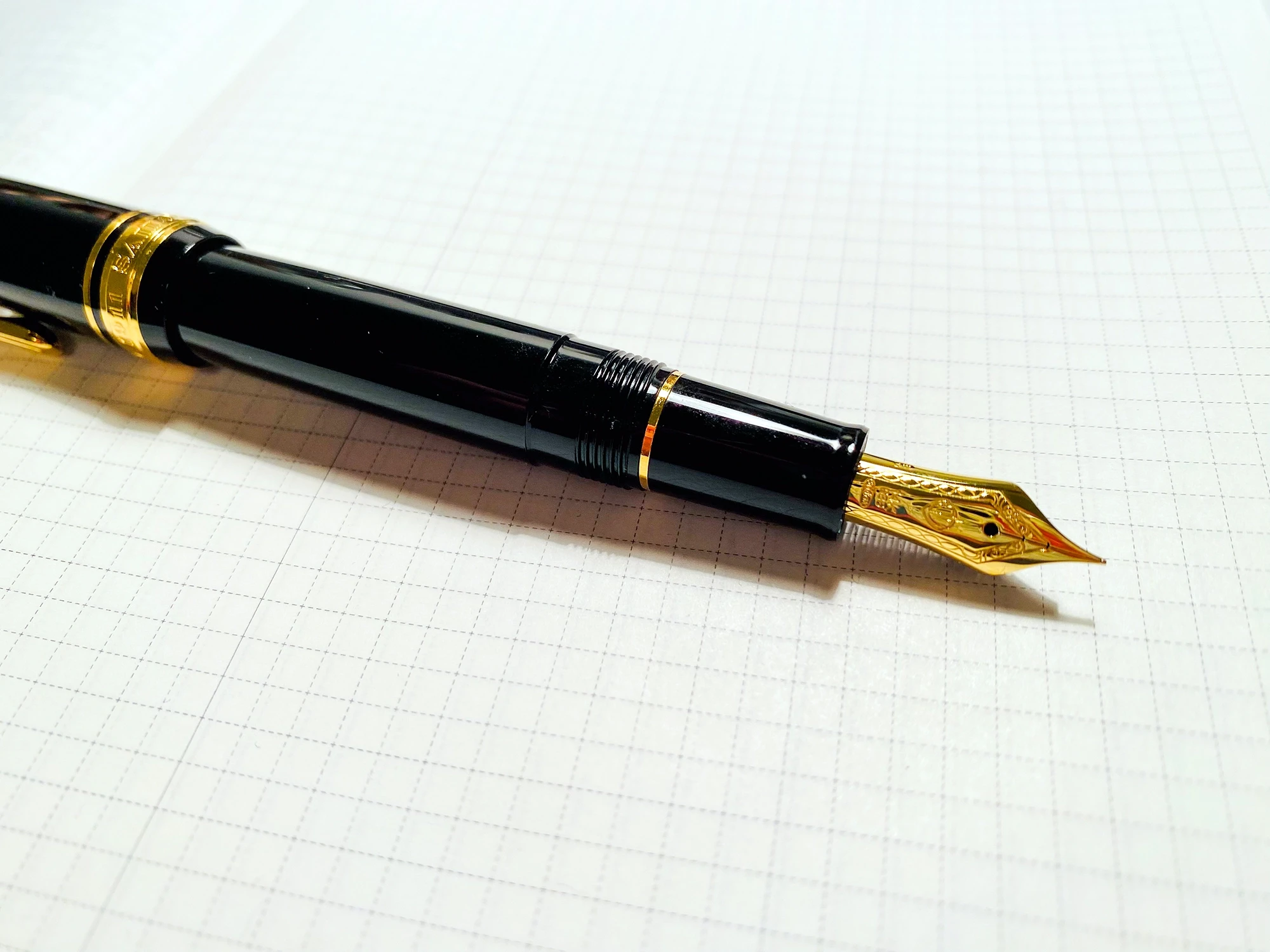A slim black fountain pen with a shiny gold nib, resting on paper with a faint graph pattern