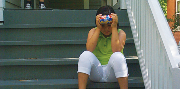 a young girl on a summer day, sitting on a stoop and holding a soda can horizontally, to cool her forehead.