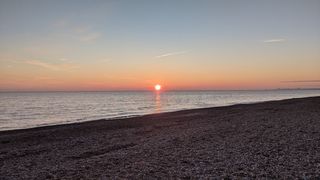 Sun setting over the sea with shingle in the foreground and the town of Worthing in the distance.