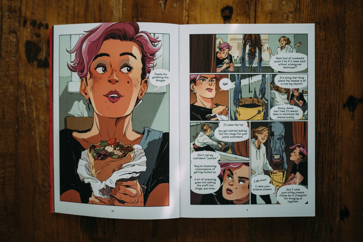 Photo of a spread from a graphic novel. One page is a close-up of a woman grabbing a bite from a wrapped sandwich. The other page depicts a casual conversation between two friends in a bathroom. Despite a dead alien hanging from the roof, they seem carefree.