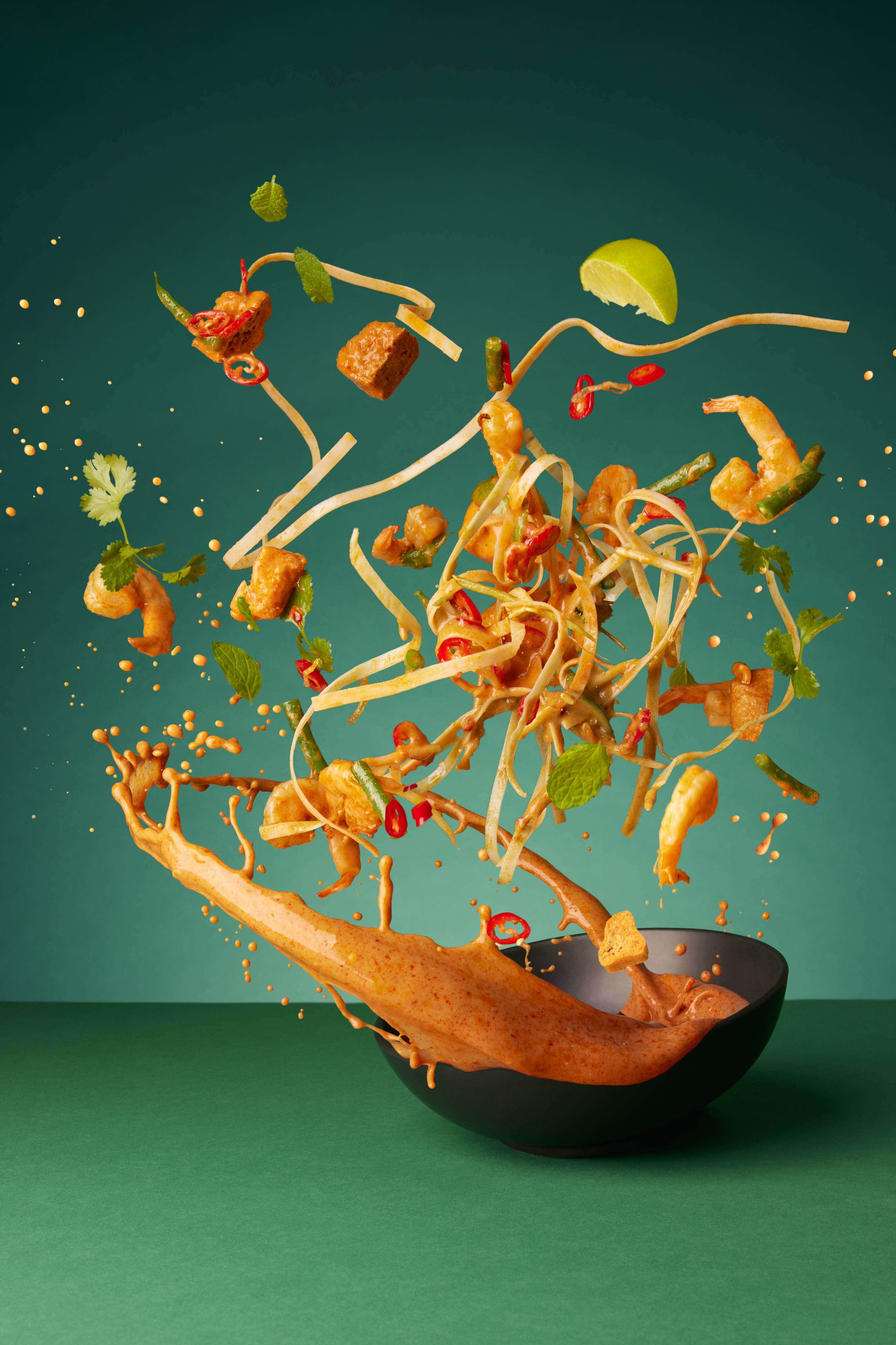 noodles flying through the air with prawns, chillis and tofu and other ingredients