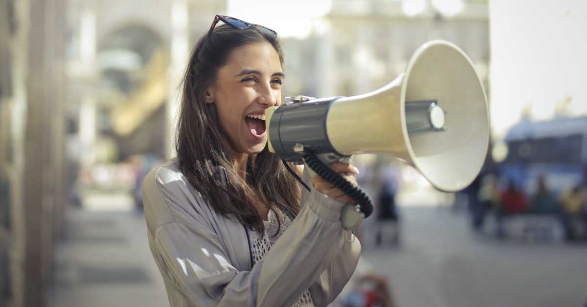 Eine junge Frau in einer grauen Jacke spricht lachend in ein Megaphon