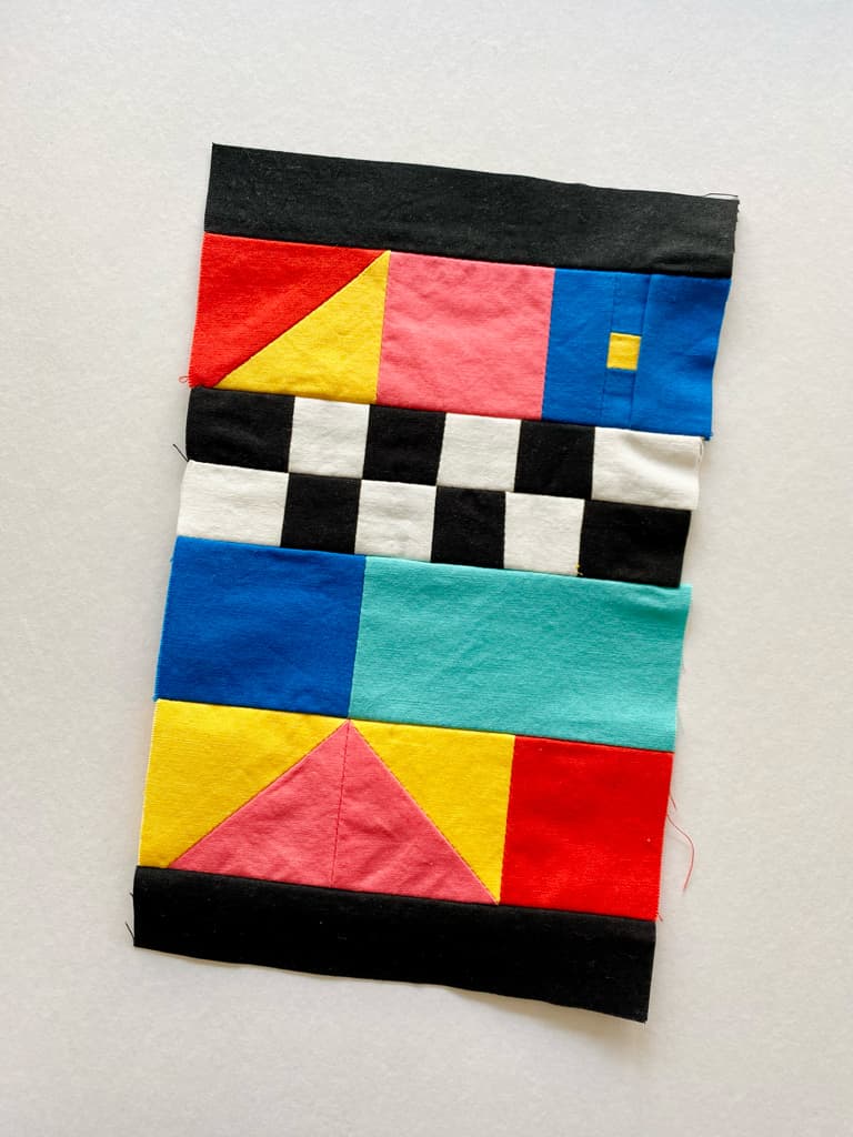A small fabric panel made of triangles, squares, and rectangles. Colors include red, yellow, pink, blue, teal, black, and white. There are black and white squares in a checkboard pattern.