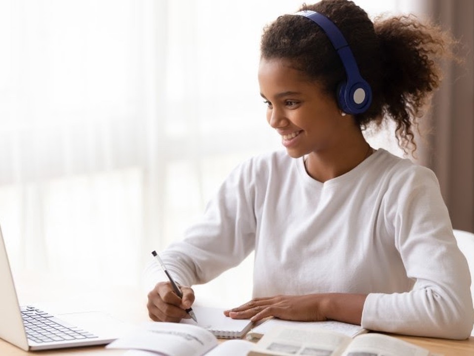 A smiling young Black student wearing headphones is taking notes while seated in front of her laptop computer.