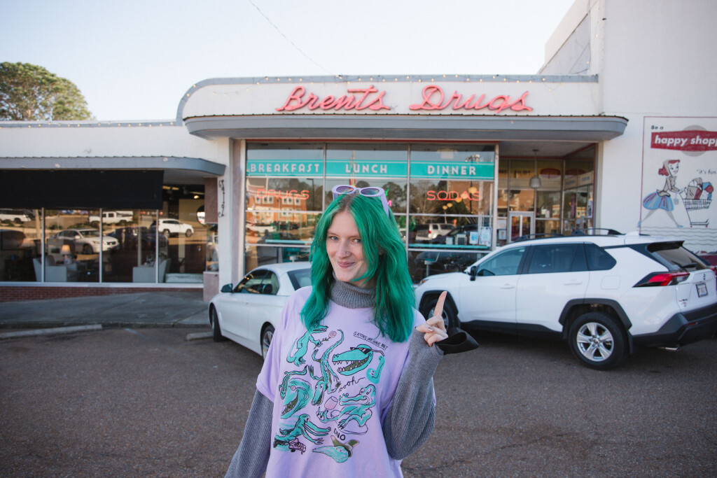 Beth's wearing a purple shirt (with drawings of gators on it) and purple sunglasses, and pointing at a sign for a diner that says Brent's Drugs