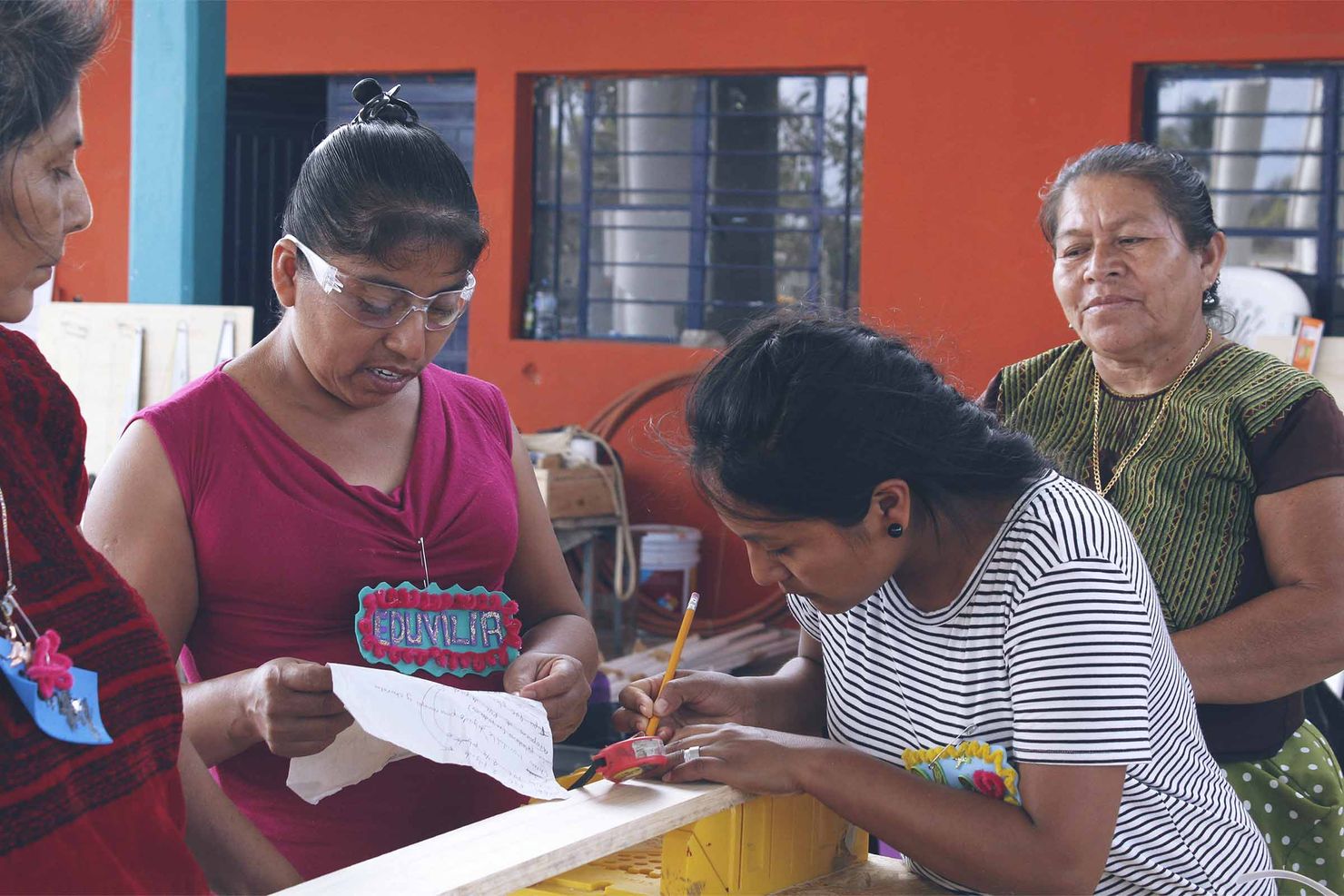 two oaxacan women collaborate to build something (one is marking a measurement on a piece of wood) while a third woman watches