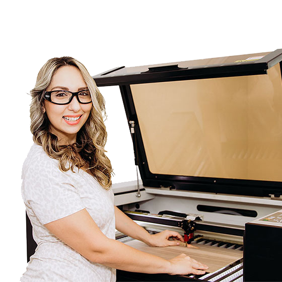 brand ambassador, Emily Caroline has long blonde wavy hair and glasses, is smiling, and standing next to an Aeon laser machine