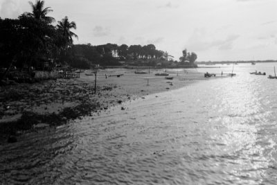 A black and white photo of the sandy seashore at Punggol Beach. A few sampans and standing wooden poles are scattered across the sand in the shallow water of the sea.