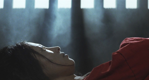 A screenshot in a smokey prison cell of a woman in orange clothes lying down staring intensely at the ceiling. From the film 'Lady Snowblood'.