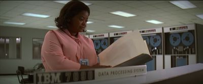 Octavia Spencer, playing NASA supervisor Dorothy Vaughan, reviews documents while standing in a computer room full of large mainframe cabinets with tape reels. A sign in the foreground reads “IBM 7090 Data Processing System.”