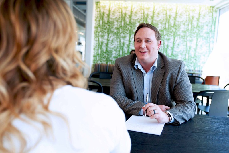 Man in a grey blazer and light blue shirt talks with a colleague at a table ready to take notes