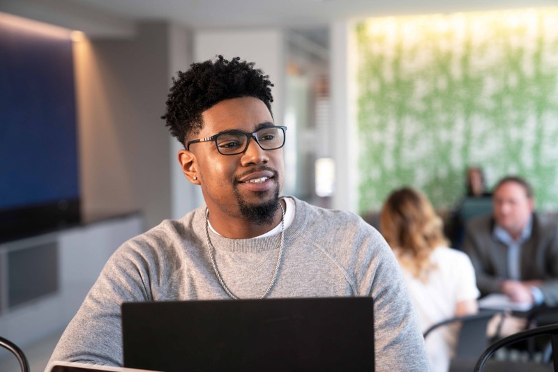 Man in grey sweatshirt and glasses looks up from his laptop as co-workers chat behind him