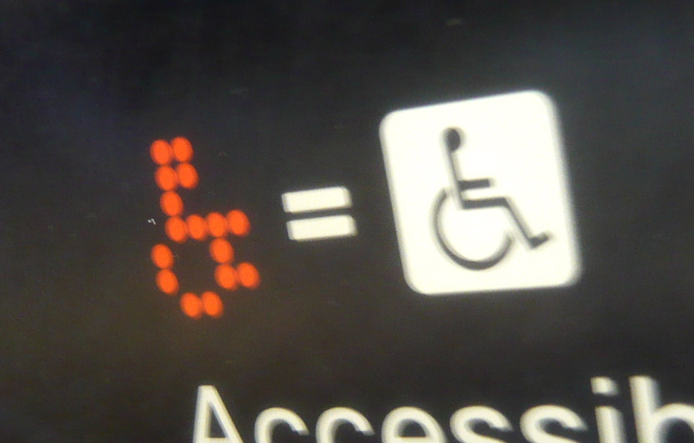 Image: a digital dot matrix wheelchair on a screen, followed by an equal symbol, and the International Symbol of Access. Below it is a partially obscured printed “accessible.” Credit: Jason Eppink on Flickr.