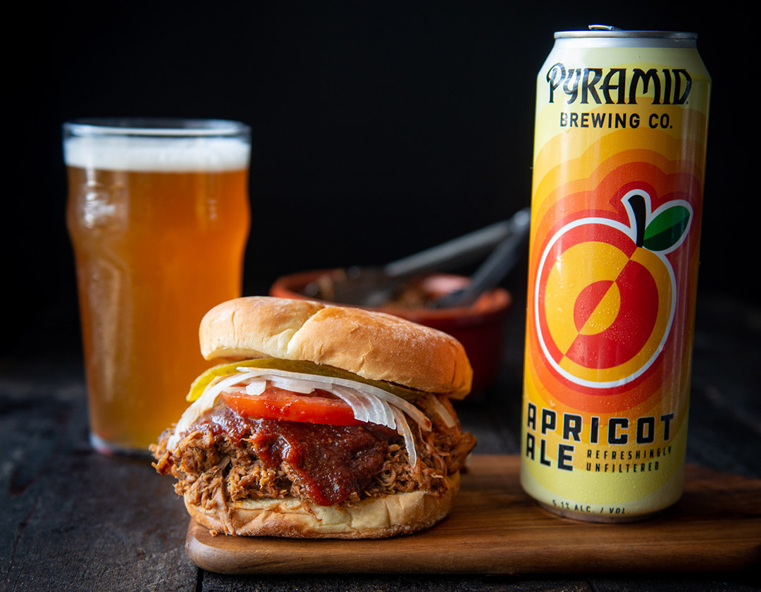Pulled pork sandwhich on a cutting board next to a can of Apricot Ale