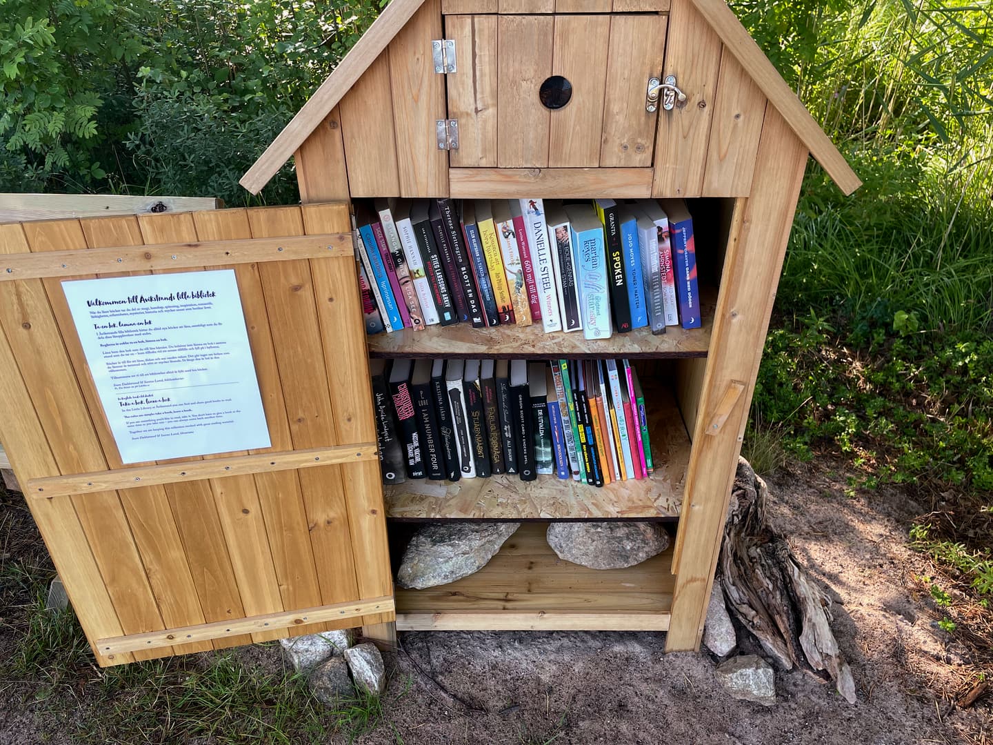 A tiny shed at a beach with the door opened. Inside there are two shelves filled with books.
