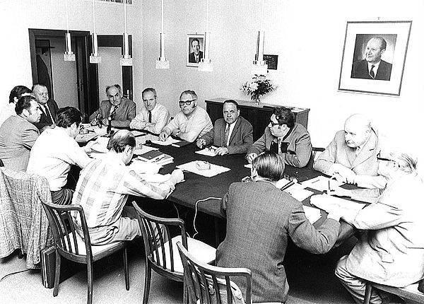 Altes schwarz-weiss Foto von 12 Männern in Anzügen die rund um einen Besprechungstisch sitzen