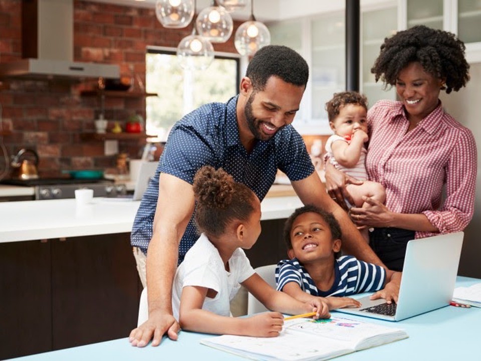 Smiling parents help their young children with homework at the kitchen table.