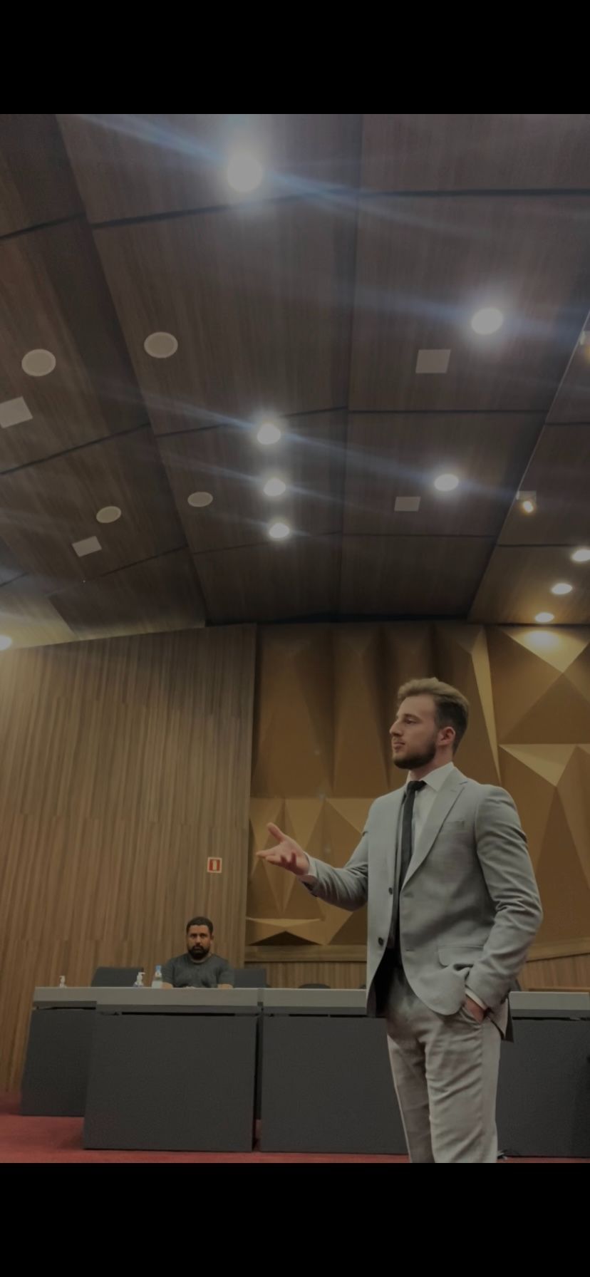 Foto de Gustavo durante audiência. É um homem branco, loiro, vestindo um terno cinza, gesticulando a mão enquanto fala.