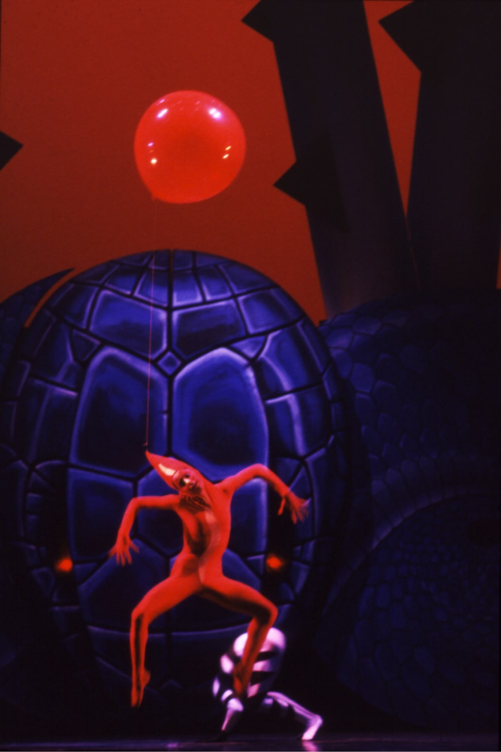 Dancer in red leotard and pointy hat leaps with red balloon in front of ominous snake head against red sky.
