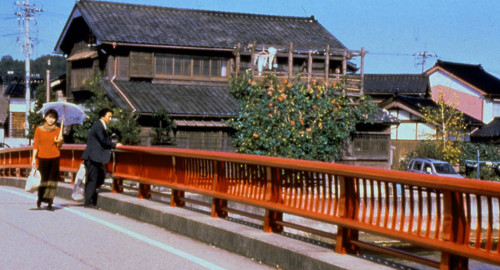 A screenshot from the film 'Warm Water Under a Red Bridge' showing an old Japanese house and bridge with a red vintage fence stretching across the screen.
