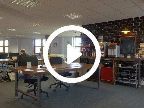 Casual shared office workspace containing long wooden computer desks and black office chairs next to beverage station Google Meet background