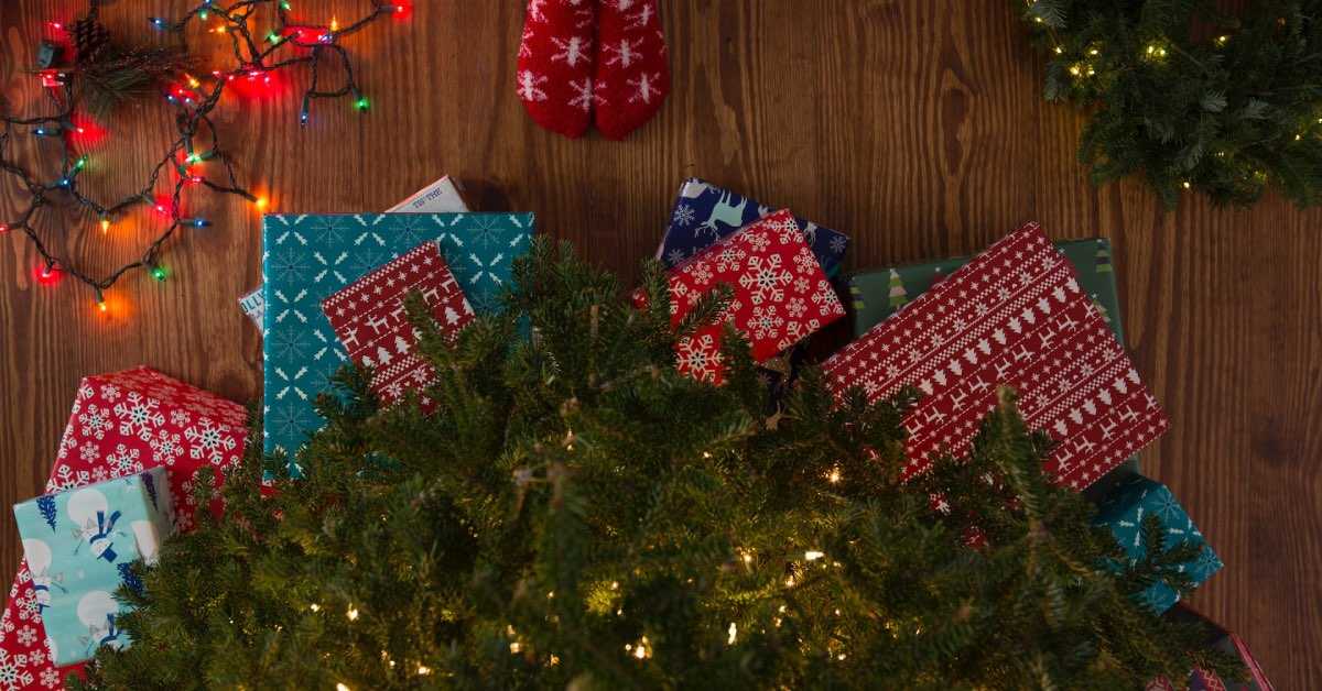 Blick auf den Boden unterhalb eines Weihnachtsbaums, zu sehen sind Geschenke, eine Lichterkette und Füße in Weihnachtssocken