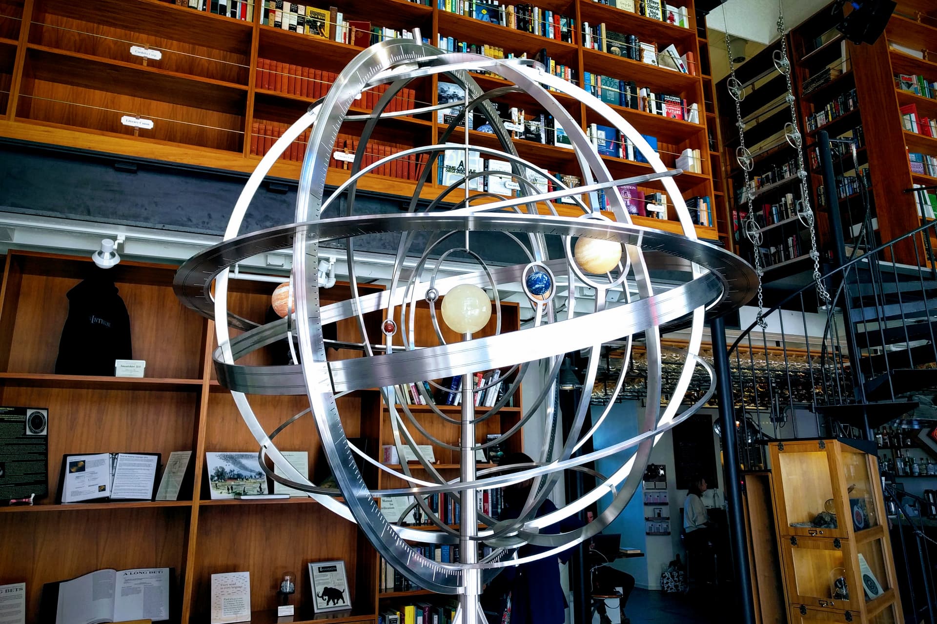 A spherical orrery made out of polished steal and stone at The Interval in San Francisco. Behind it are two floors of bookshelves and a spiral iron staircase.