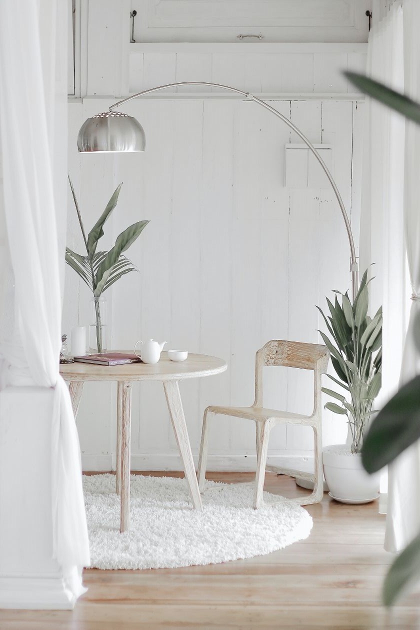 Ein Tisch und ein Stuhl aus hellem Holz auf einem runden weißen Teppich. Drum herum ein paar Pflanzen und eine Lampe.