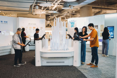 A photo of the Singaporium exhibition. Walls feature reproductions of books and photos. In the foreground, there is a sculpture of frosted acrylic tubes that light up. Visitors are exploring the content and the sculpture.