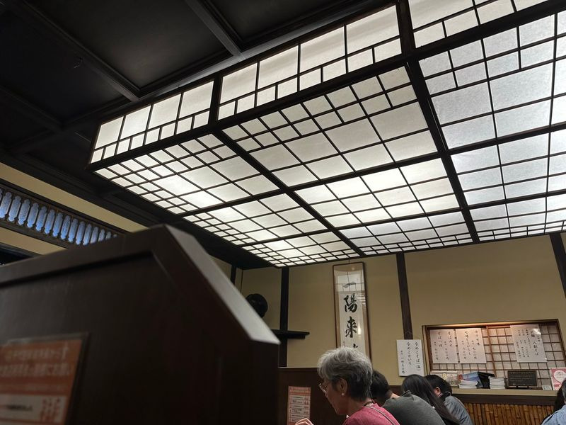 The ceiling lamp of the restaurant. It’s large and rectangular, dominating the ceiling. It’s composed of a square, dark wooden lattice and white paper reminiscent of traditional Japanese sliding doors. Some patrons can be seen below.