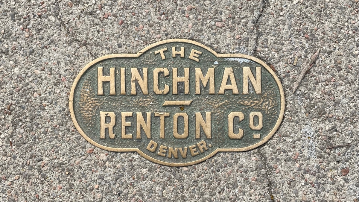 A bright brass seal reads 'The Hinchman-Renton Co., Denver' is set in an aging, pebbled sidewalk