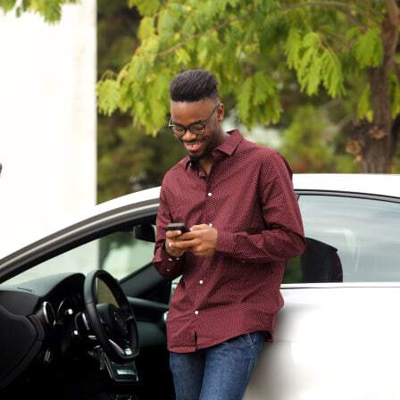 Man in red shirt leans against car looking at smartphone.