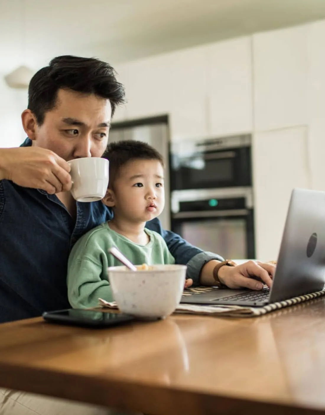 man of color sipping coffee while holding his son in his lap and working on a laptop