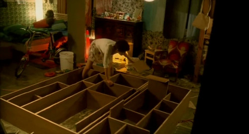 A screenshot from the film 'About Love' of A-su bending down to fix the massive bookshelf that is on her partment floor.