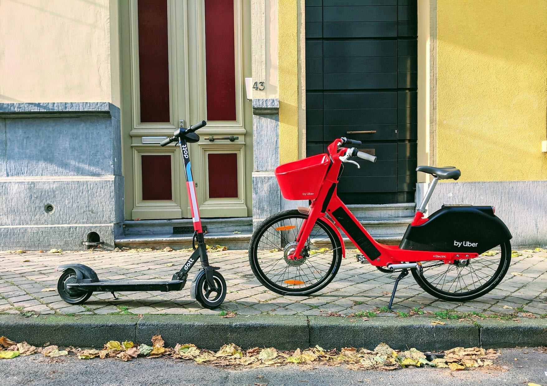 A red and blue kick-scooter and a red free-floating bike with a basket faced against each other in the middle of an empty street.