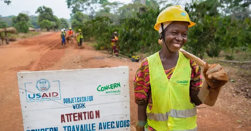 Smiling woman with hard hat and pick axe, standing on road beside a sign
