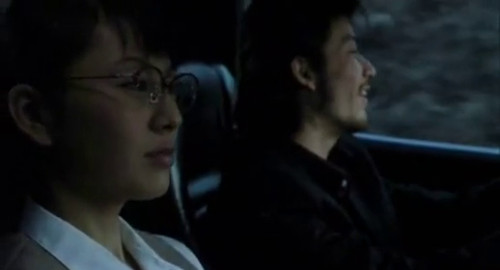 A screenshot of a worried woman in glasses driving a car with an energetic young man in the passengers seat. From the movie 'Shark Skin Man and Peach Hip Girl'.