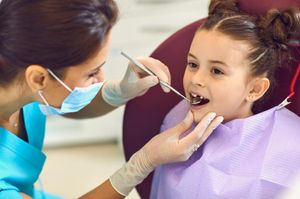 A young girl in a dentist's chair. A dental professional wearing a mask and gloves is examining her mouth with a tool.