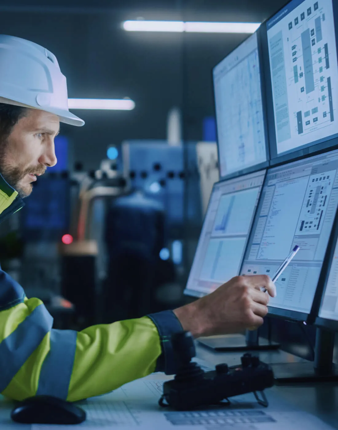 bearded man wearing a hardhat and safety jacket and is pointing at one of many monitors at a desk