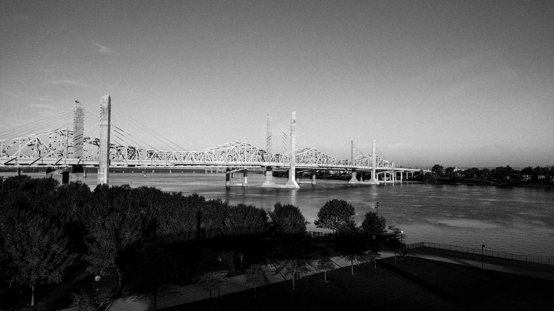 A black-and-white photograph of a brutalist suspension bridge of white steel and concrete pylons crossing the Ohio River.